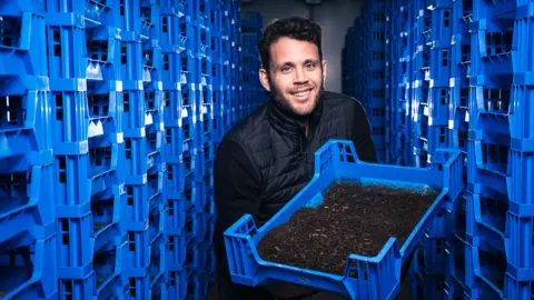 Flybox Larry Kotch тримає синій контейнер, повний личинок мухи
