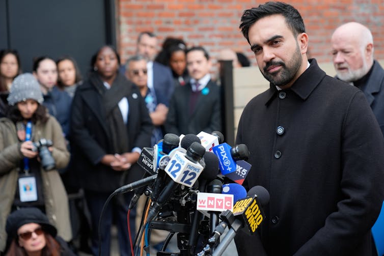 Чоловік у краватці та піджаку з багатьма мікрофонами перед ним.